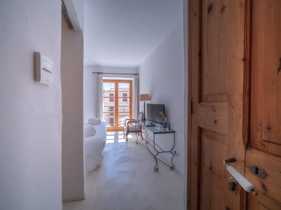 Un couloir avec une porte en bois, un lit, un bureau avec une lampe et une fenêtre donnant sur des immeubles.