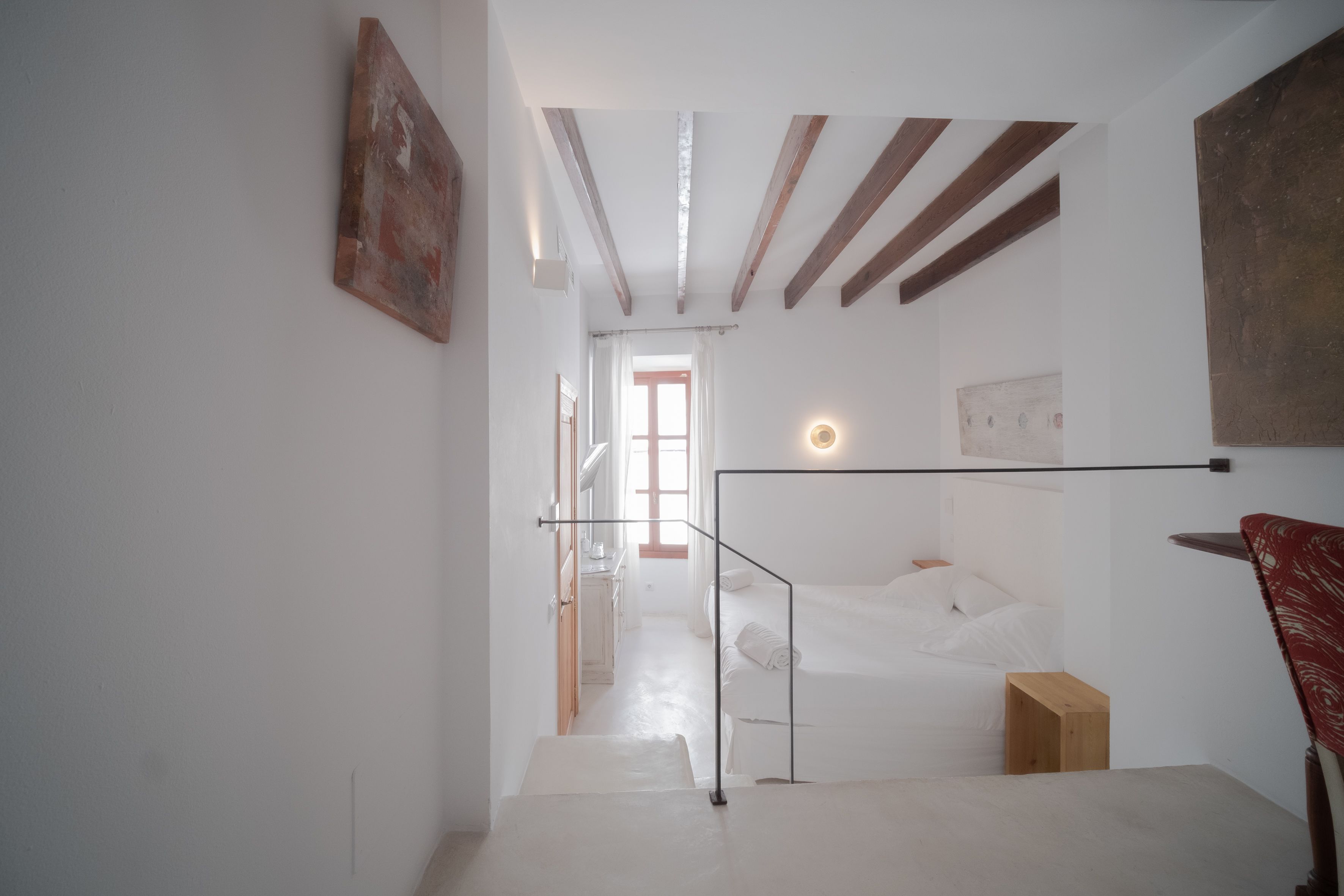 Habitación de hotel moderna con diseño minimalista, que incluye una cama, un taburete de madera y una silla roja.
