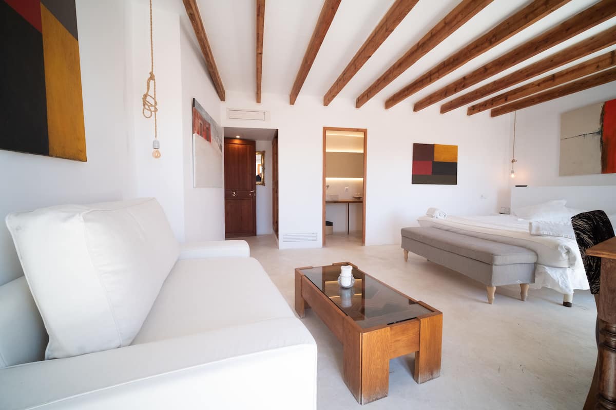 Ein modernes Wohnzimmer mit einem weißen Sofa, einem Holztisch und einer grauen Bank. Die Holzbalkendecke ist sichtbar, und eine Pendelleuchte hängt von der Decke.