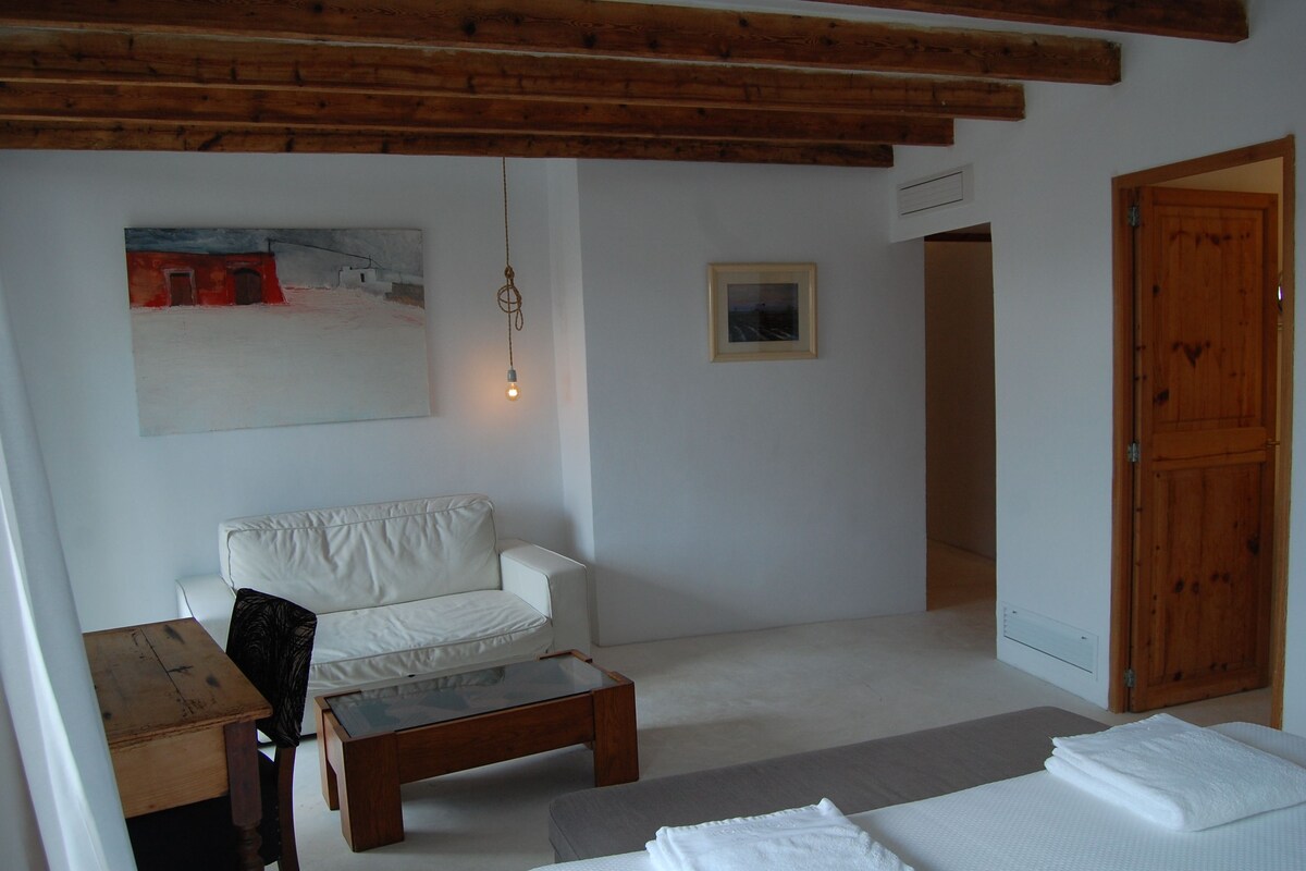 A room with a white sofa, a wooden coffee table, a bed with white linens, a wooden door, and a painting on the wall.