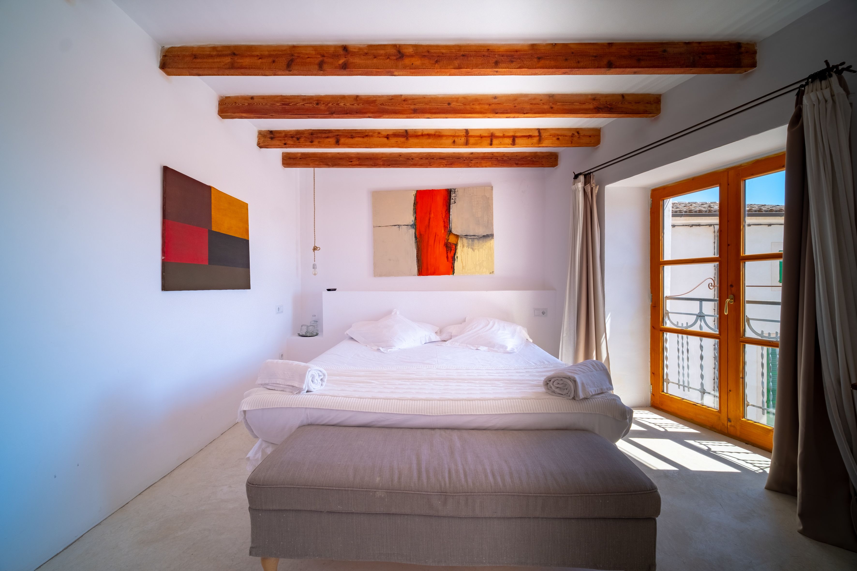 A bedroom with a bed, a bench, wooden beams, a window, and abstract art on the wall.