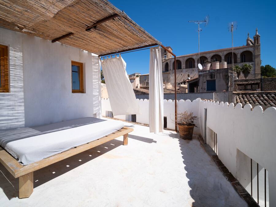 Un balcó amb un llit, parets blanques, un sostre de fusta i vista d'un edifici històric amb arcs.