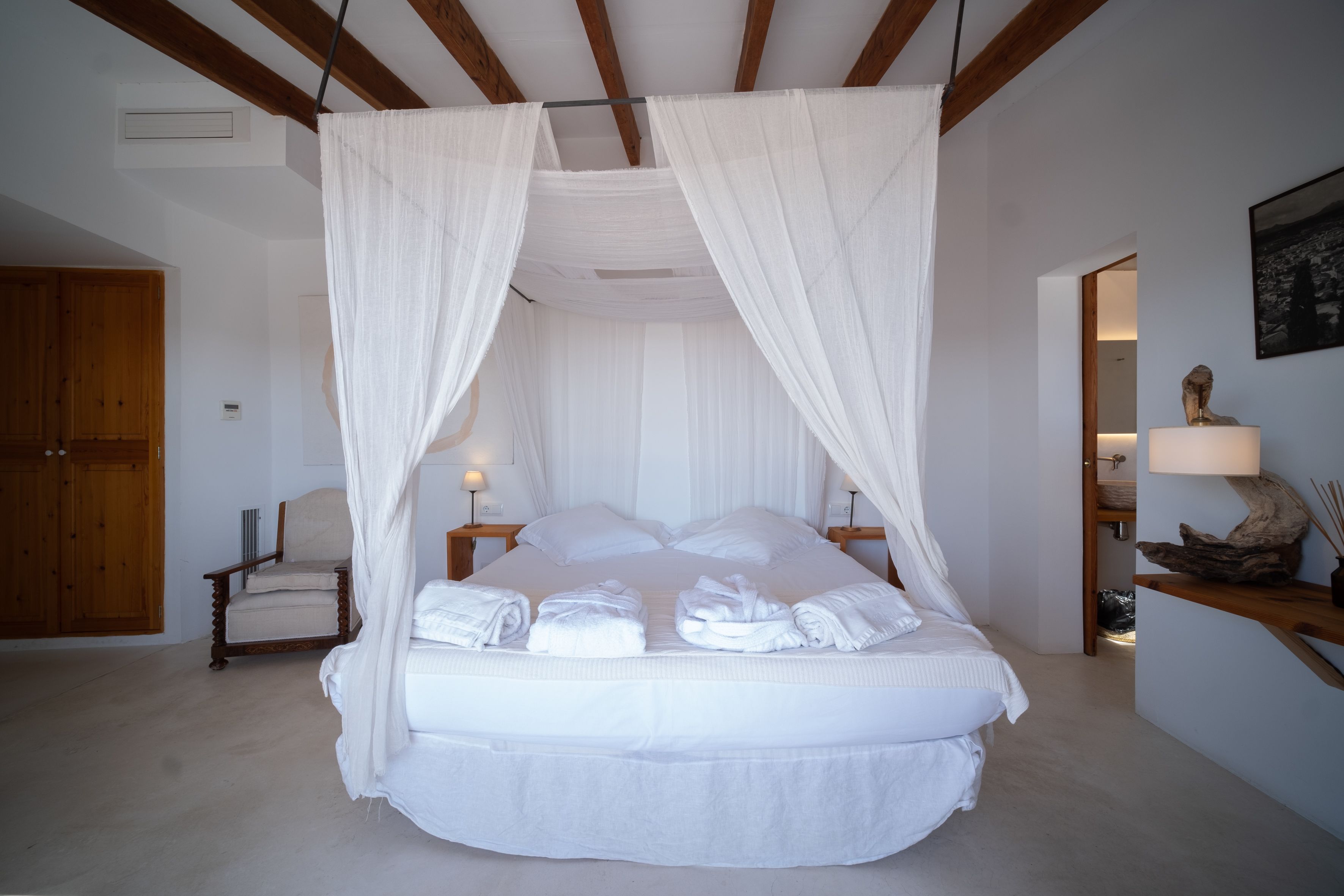 Una cama con dosel y cuatro columnas con ropa de cama y toallas blancas, una silla de madera, una mesita de noche con una lámpara y una estantería de madera con objetos decorativos.