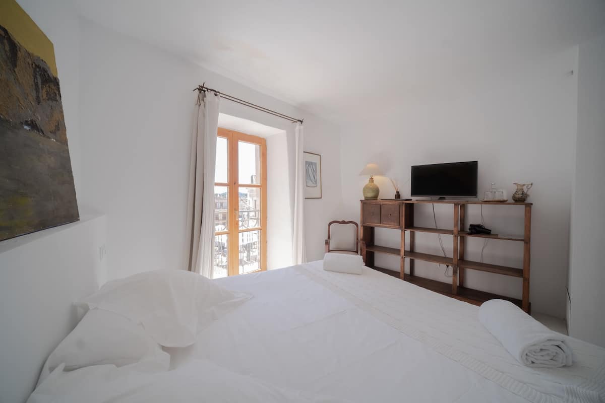 Ein Schlafzimmer mit Bett, Fernseher auf einem Holzboden, einem Fenster mit Vorhängen und einem Gemälde an der Wand.