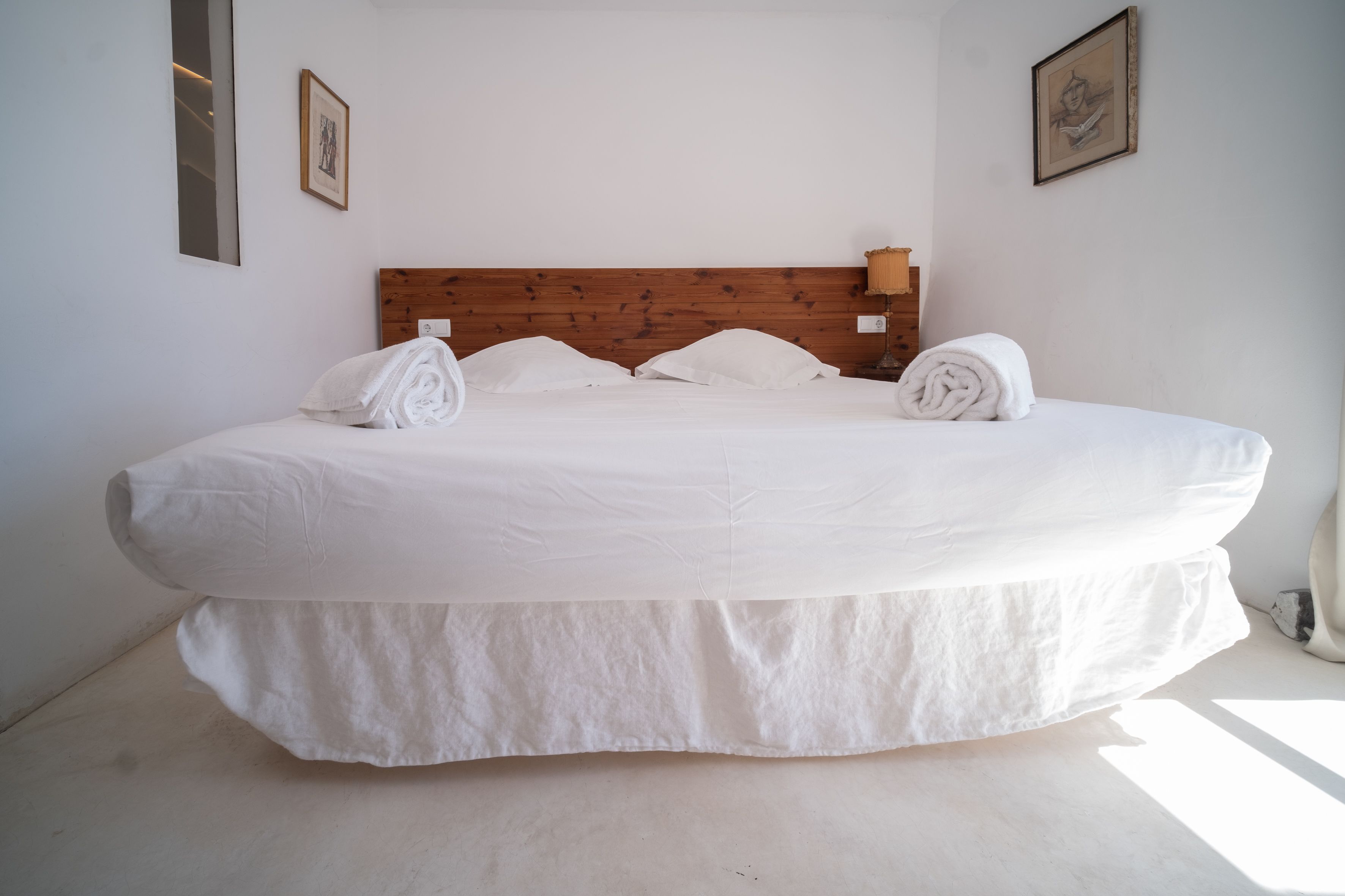 A large bed with white linens and a wooden headboard in a bright room with white walls.