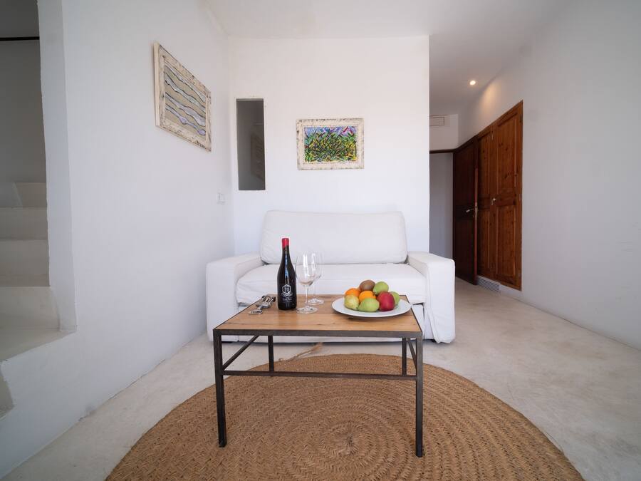 Un salón con un sofá blanco, una mesa de centro con una botella y vasos, y un frutero.