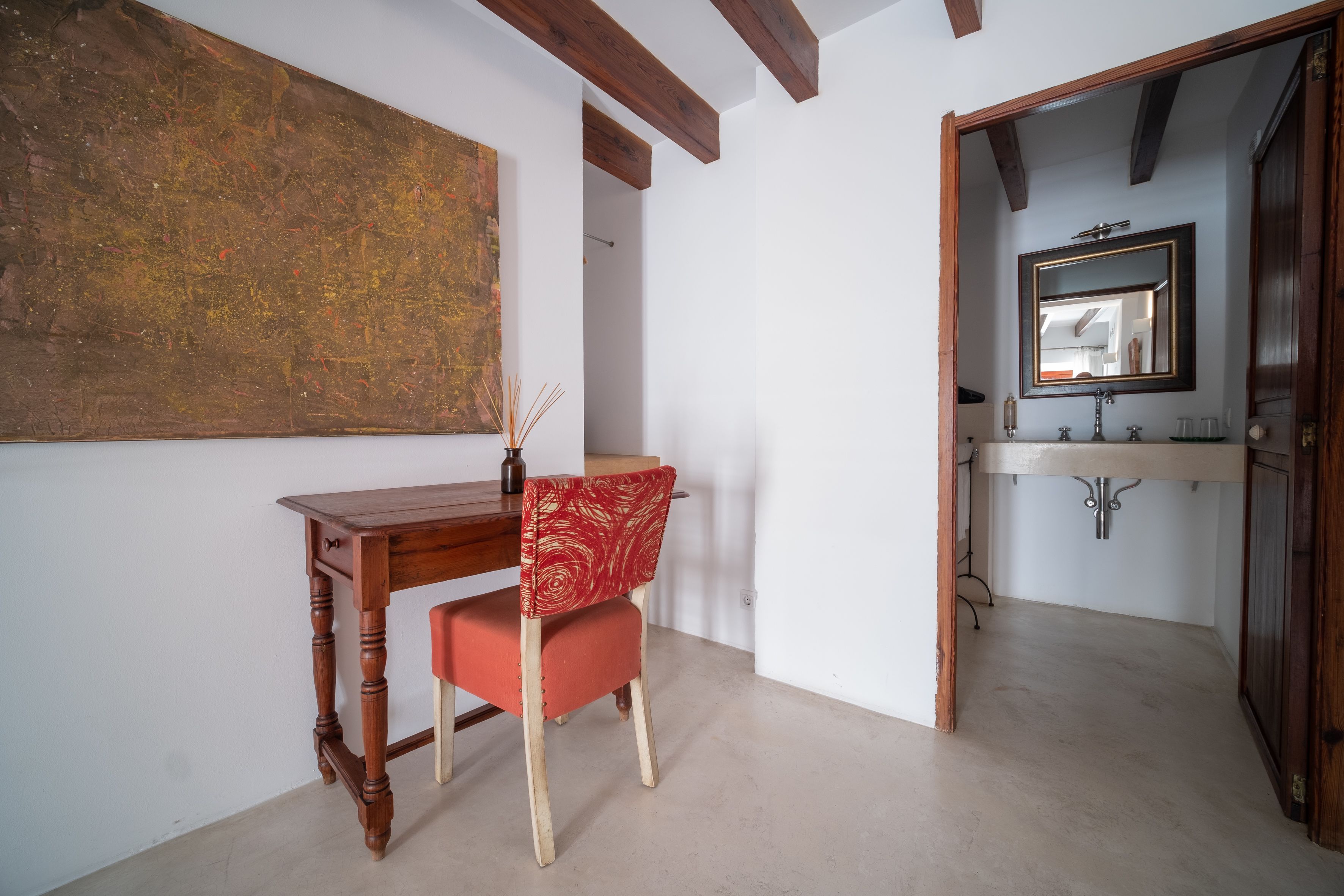Una habitación con un escritorio de madera, una silla roja, un cuadro abstracto grande y un baño con espejo y lavabo.