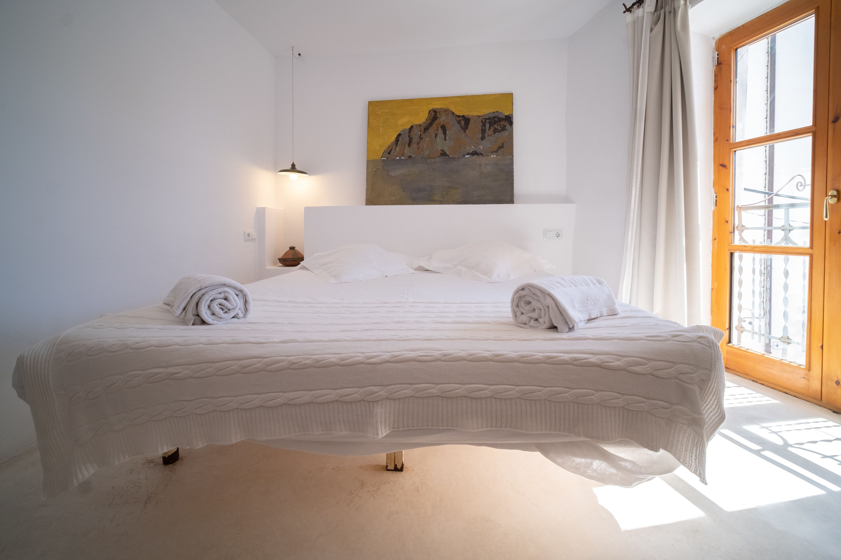 Een keurig opgemaakt bed met witte lakens en een schilderij van een berg aan de muur.