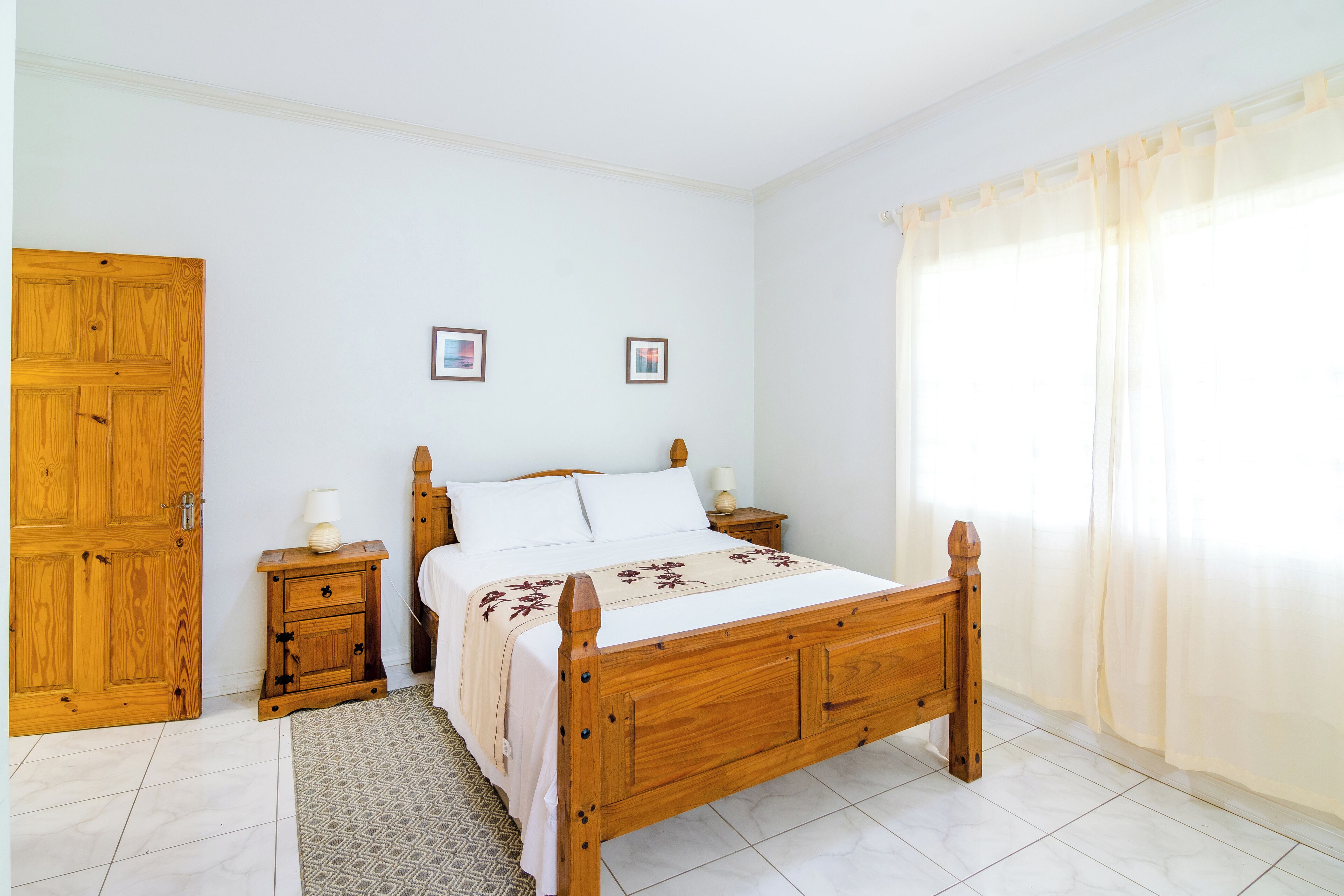 A bedroom with a wooden bed, a nightstand, a wooden wardrobe, and a window with sheer curtains.