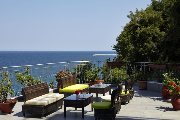 Un área de estar al aire libre con mobiliario de mimbre, plantas en macetas y vista al mar.