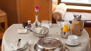 Mesa de jantar posta com talheres, pratos, uma tigela, um jarro, um vaso com uma rosa e um pratinho com condimentos.
