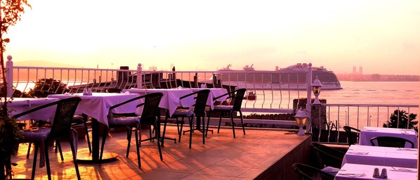 An outdoor dining area with tables and chairs, overlooking a body of water with a cruise ship in the background.