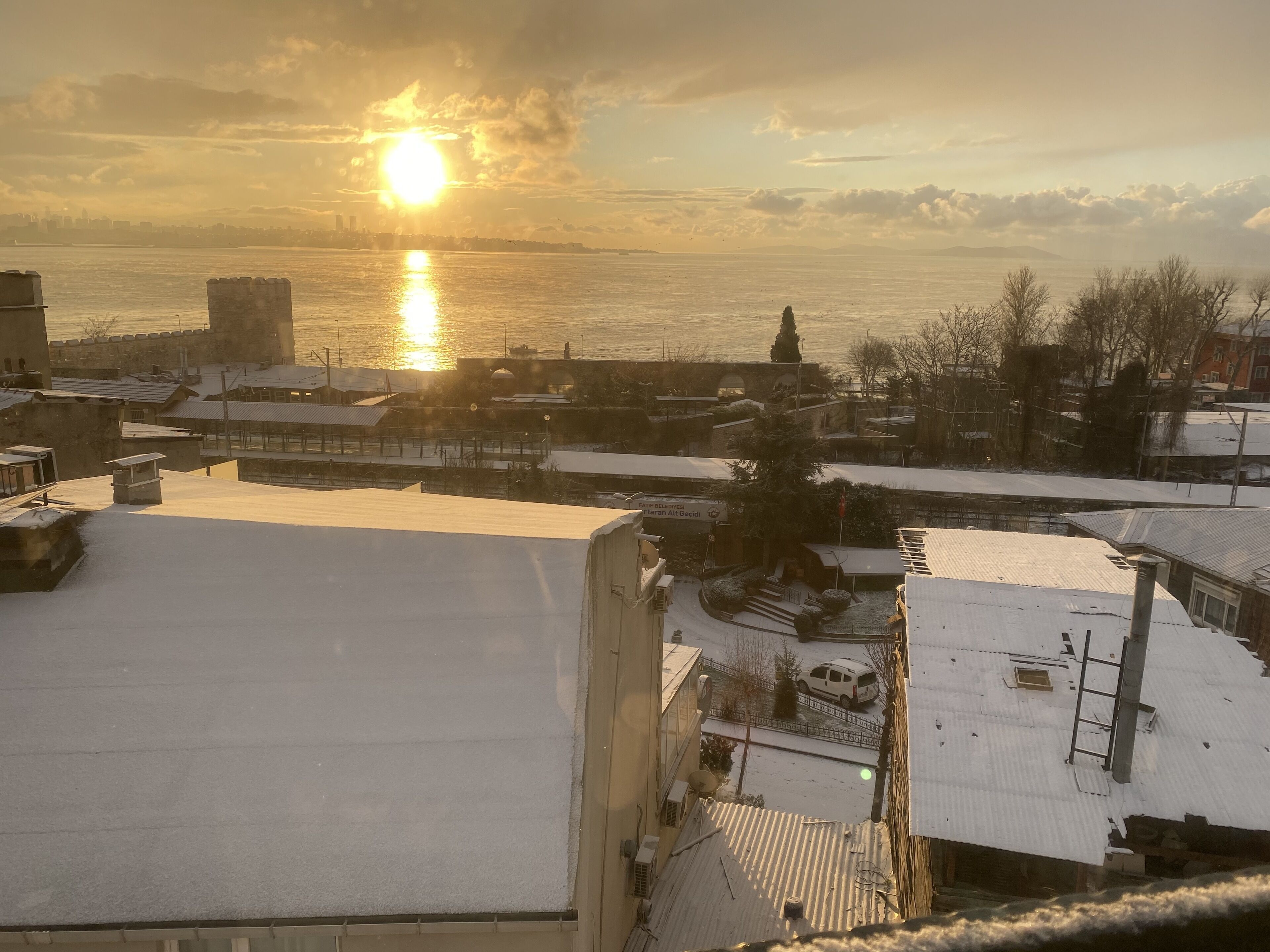 A snowy urban landscape with buildings and a body of water during sunset.