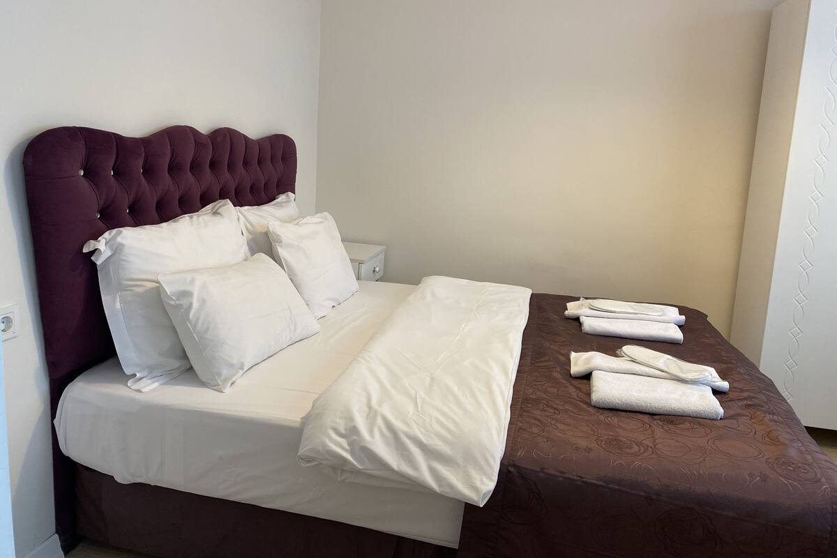 A neatly made bed with white linens and a brown headboard, a bedside table with a lamp, and folded towels on the bed.