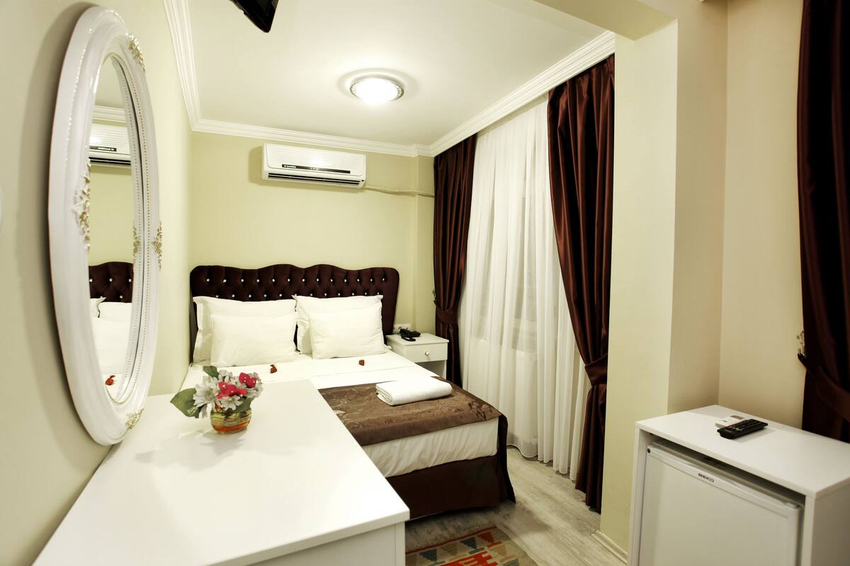 A hotel room with a bed, a bedside table, a mirror, an air conditioning unit, a desk, and a small refrigerator.