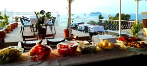 A terrace with tables and chairs, potted plants, and a view of the sea and a cruise ship.