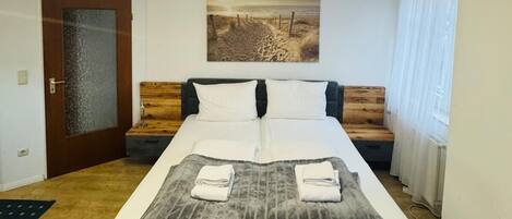 Una habitación de hotel moderna con una cama grande, cabecera de madera, un cuadro en la pared y una lámpara de techo de diseño geométrico.
