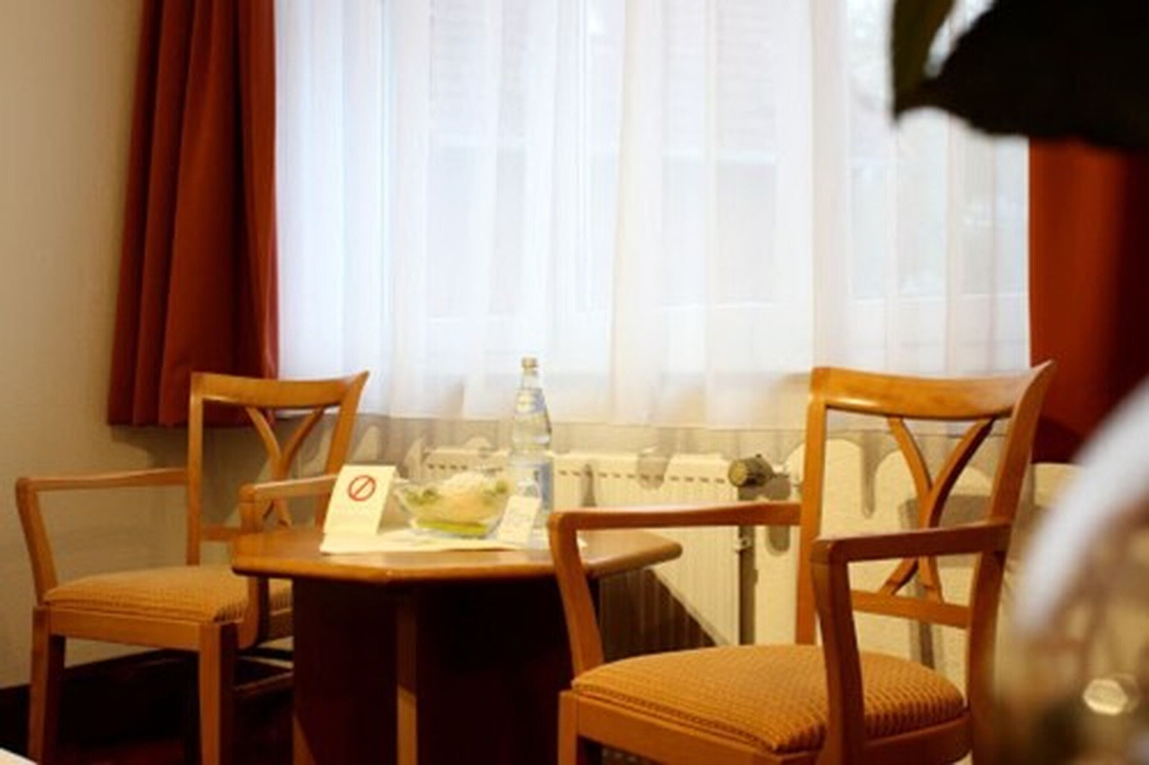 Et hotelværelse med et lille bord, to stole, et vindue med lette gardiner og en radiator.