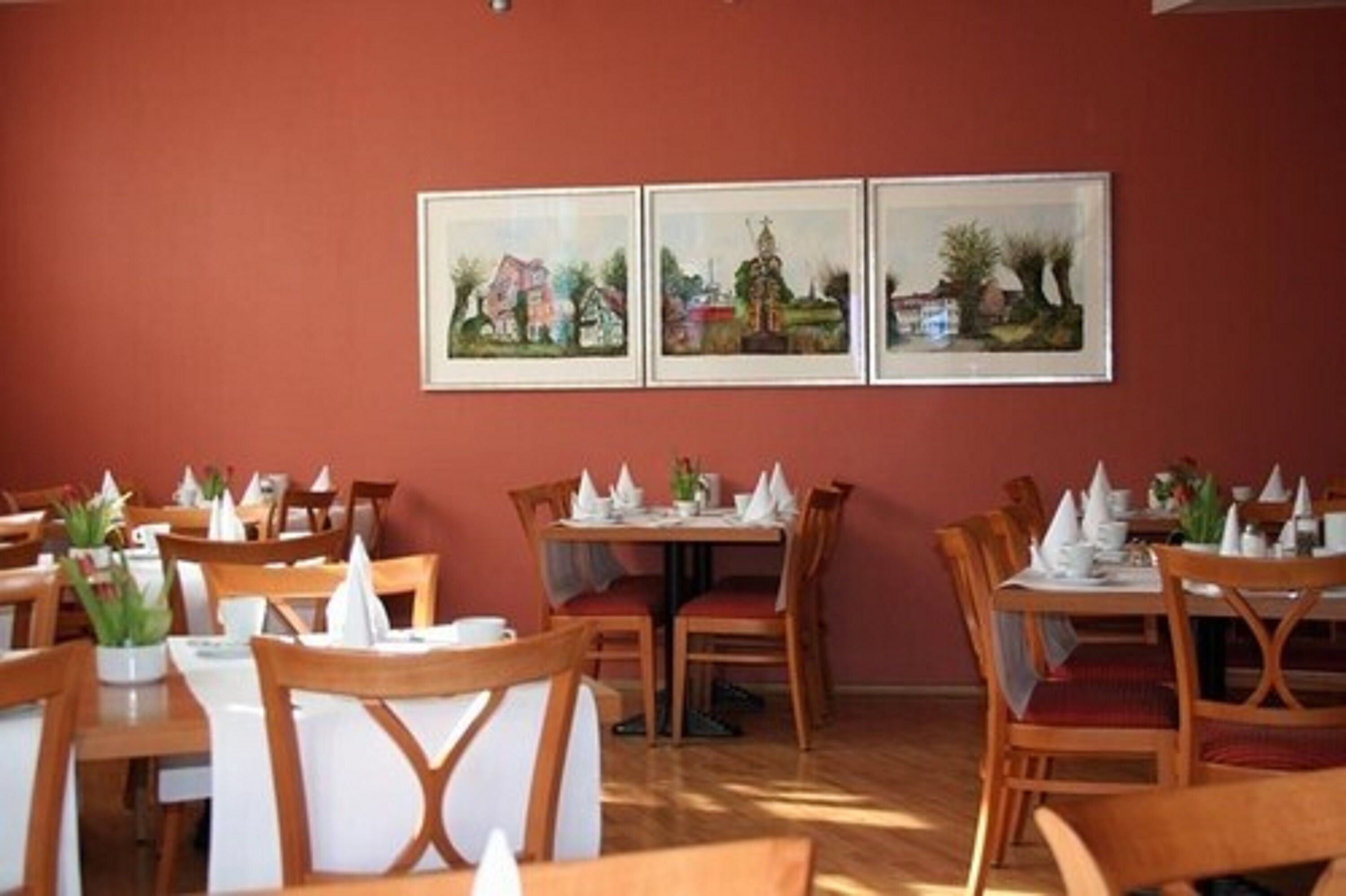 A dining area with wooden tables and chairs, white tablecloths, and napkins. There are framed pictures on the wall.