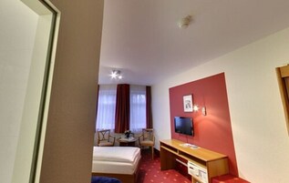 Quarto de hotel com cama, escrivaninha, cadeira, televisão e janela com cortinas.