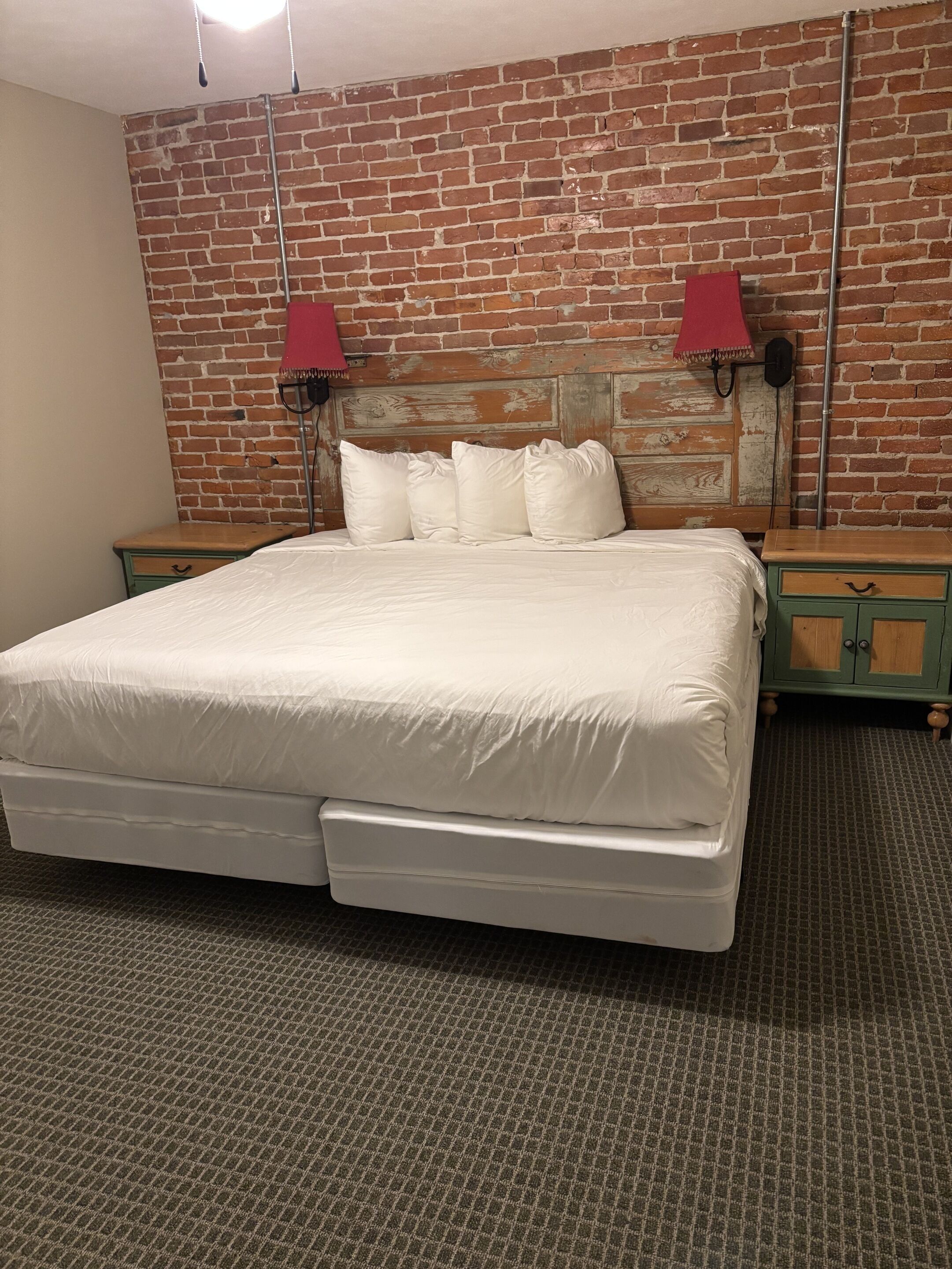 Una habitación con pared de ladrillo, una cama con ropa de cama blanca, dos mesitas de noche y un armario verde.