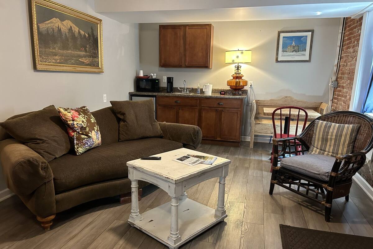 A living room with a brown sofa, a white coffee table, a wicker chair, wooden flooring, a kitchen area with wooden cabinets, a lamp, and a framed picture on the wall.