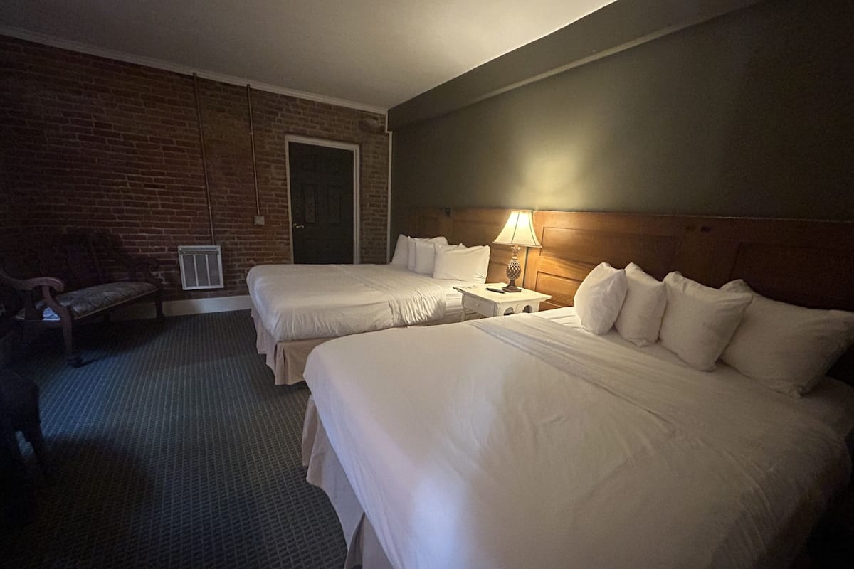 A hotel room with two beds, a bench, a lamp, and a brick wall.