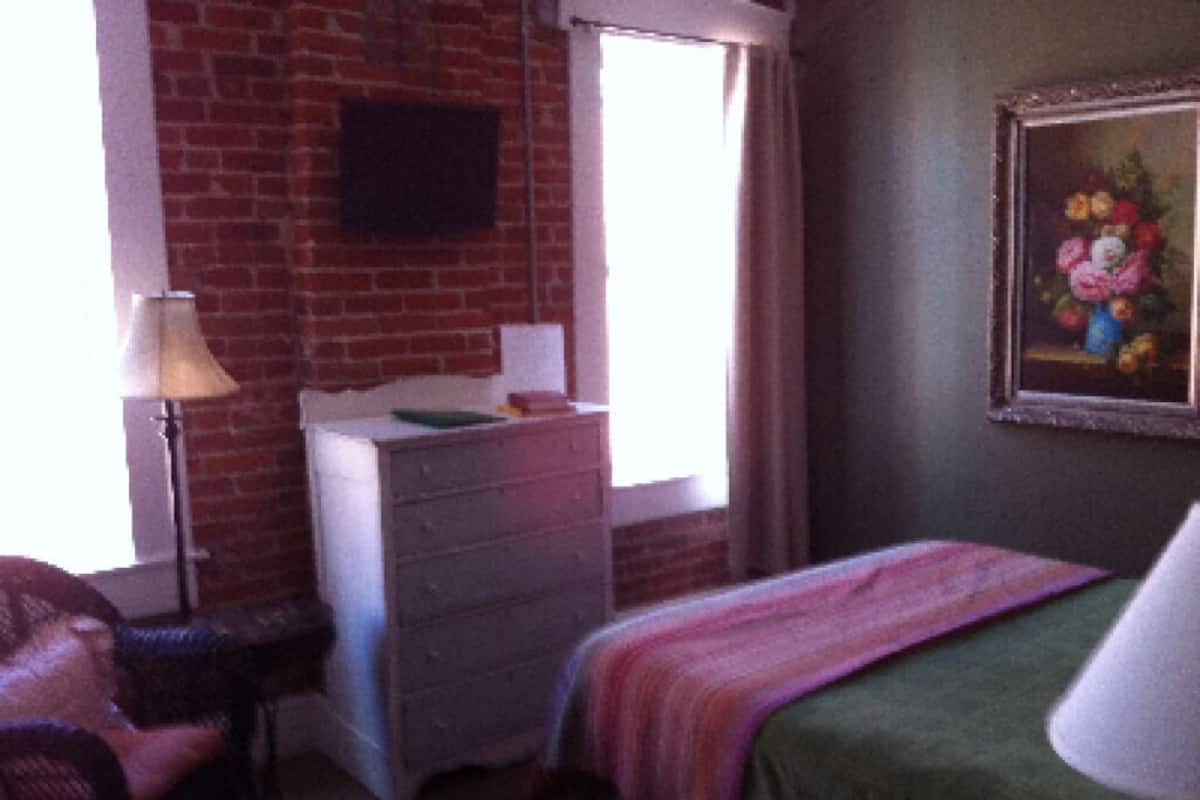 A room with a brick wall, a desk with a lamp, a bed with a green blanket, and a painting on the wall.