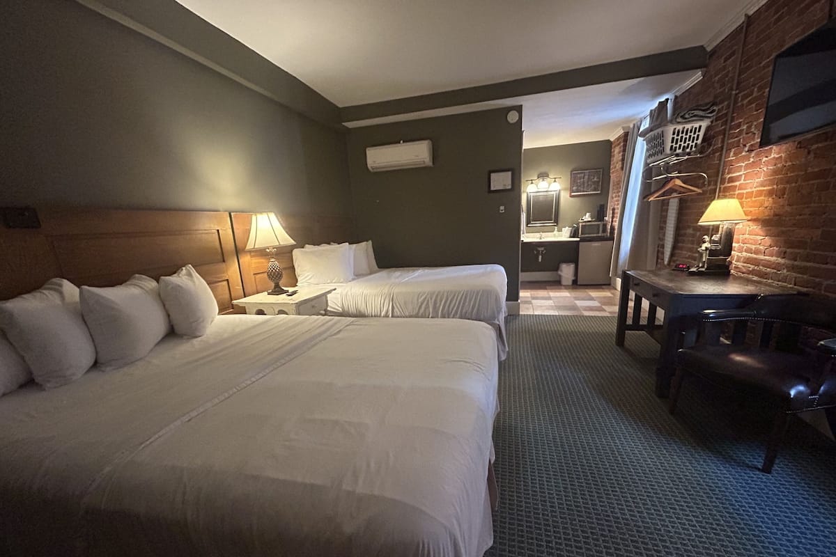 A hotel room with two beds, a desk, a chair, a lamp, a small table, and a bathroom visible in the background.