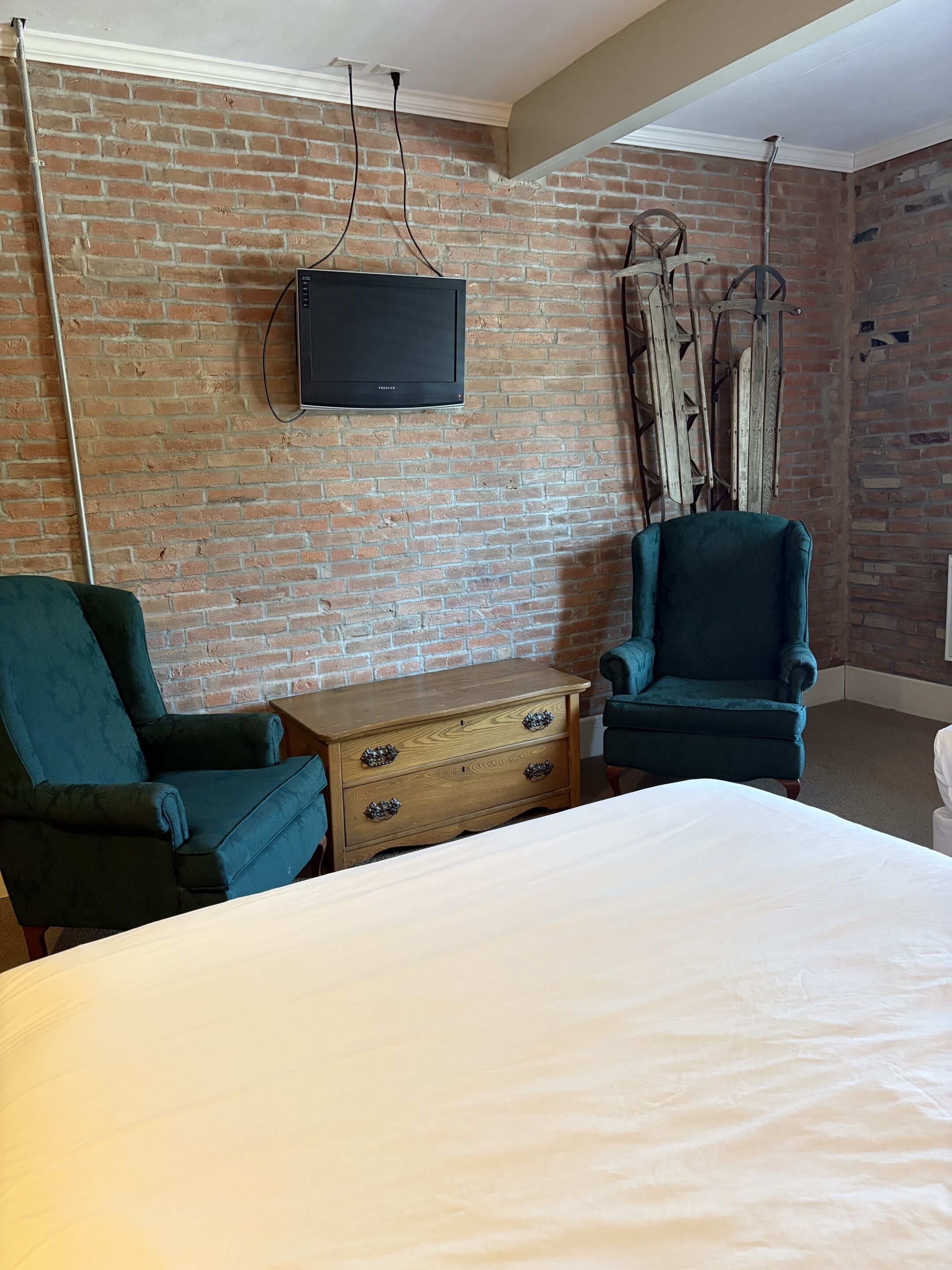 A room with a brick wall, a television mounted above a wooden dresser, two teal armchairs, and a bed with white bedding.