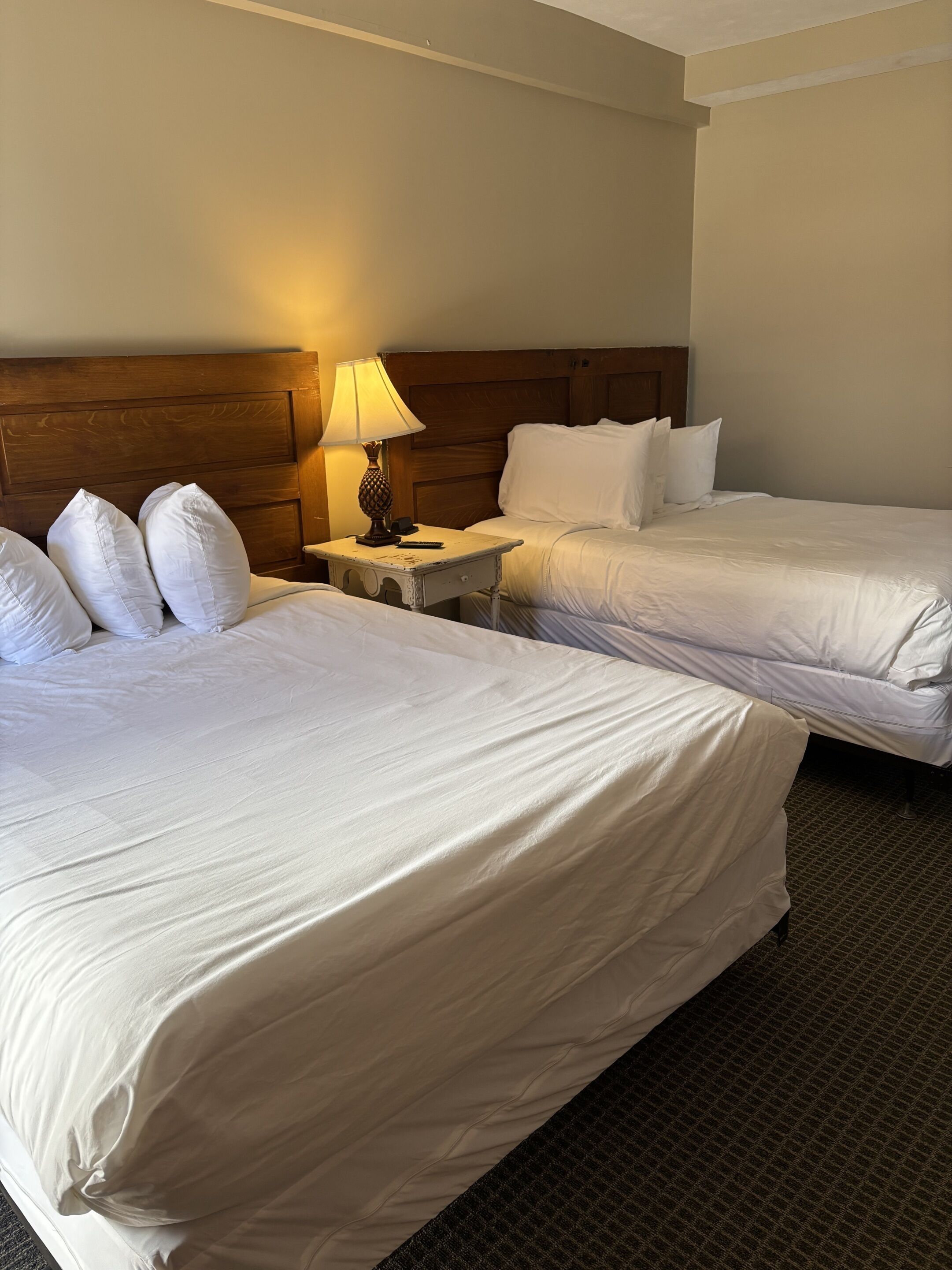 A hotel room with two beds, a wooden headboard, a nightstand with a lamp, and a carpeted floor.