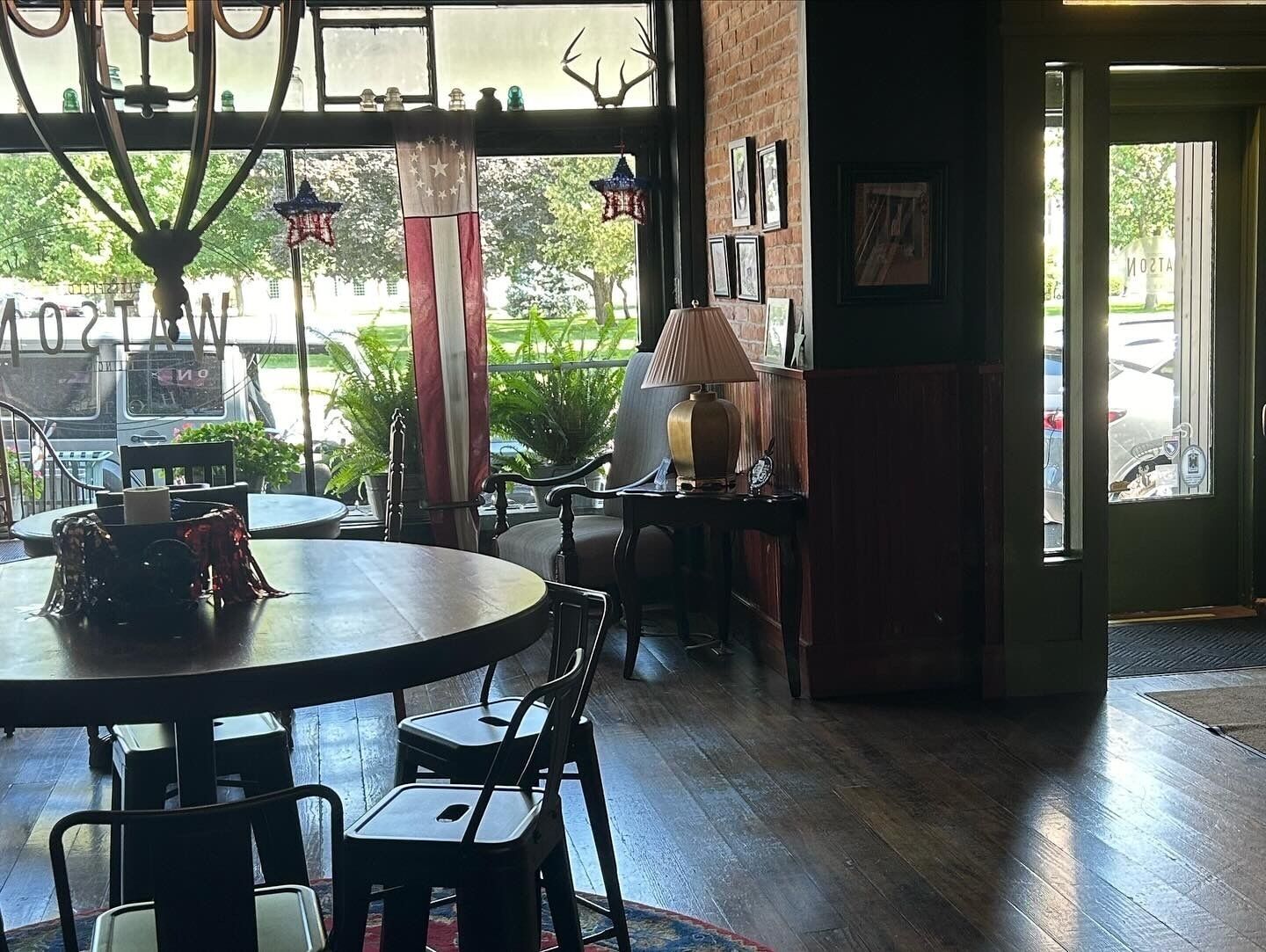 Interior acolhedor de restaurante com uma mesa redonda, cadeiras, um abajur e vista para a rua através de grandes janelas.