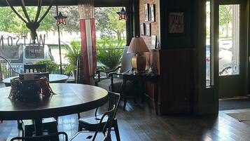 Interior acolhedor de restaurante com uma mesa redonda, cadeiras, um abajur e vista para a rua através de grandes janelas.