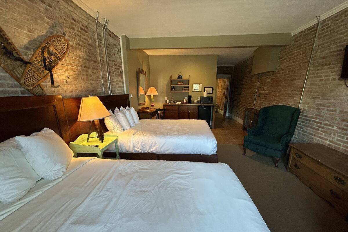 A hotel room with two beds, a desk, a chair, a TV, and a kitchenette.