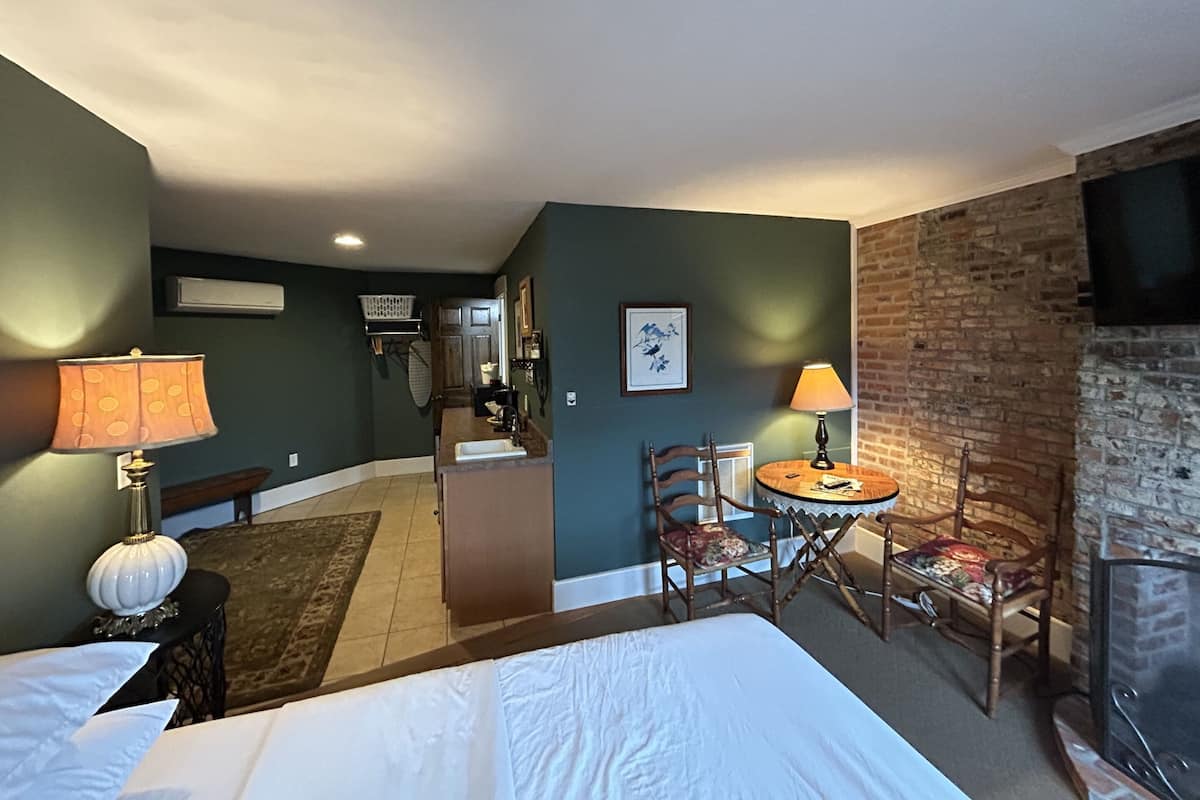 A hotel room with a bed, a dining table with chairs, a fireplace, and a kitchen area in the background.