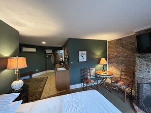 A hotel room with a bed, a dining table with chairs, a fireplace, and a kitchen area in the background.