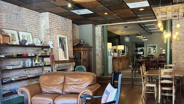 A cozy living area with a leather sofa, a wooden coffee table, and a patterned armchair. A brick wall and wooden shelves with various items are visible. - William Watson Hotel (Pittsfield)