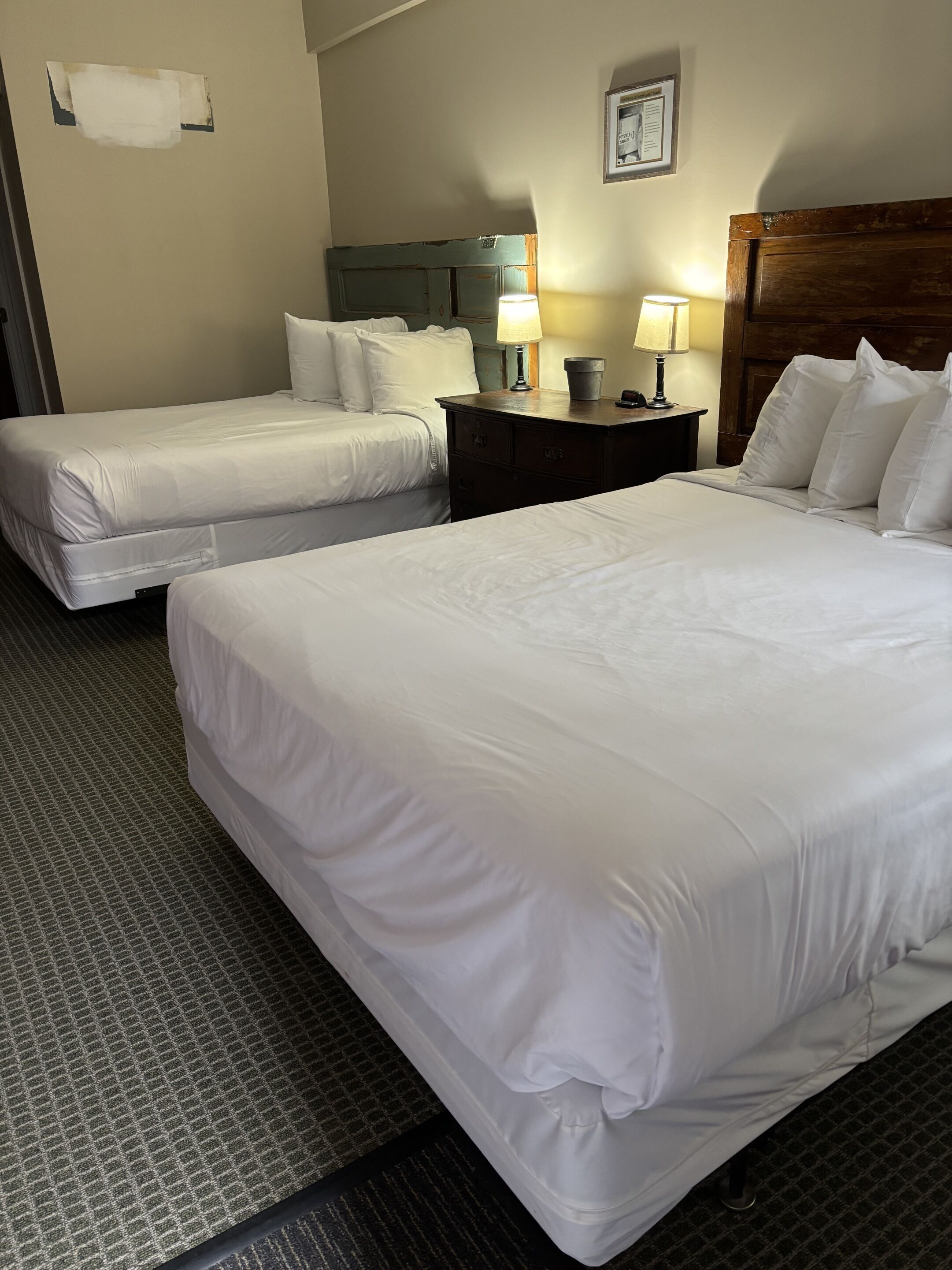 Een hotelkamer met twee bedden, een houten hoofdbord, een nachtkastje met een lamp en een lijst met een afbeelding aan de muur.