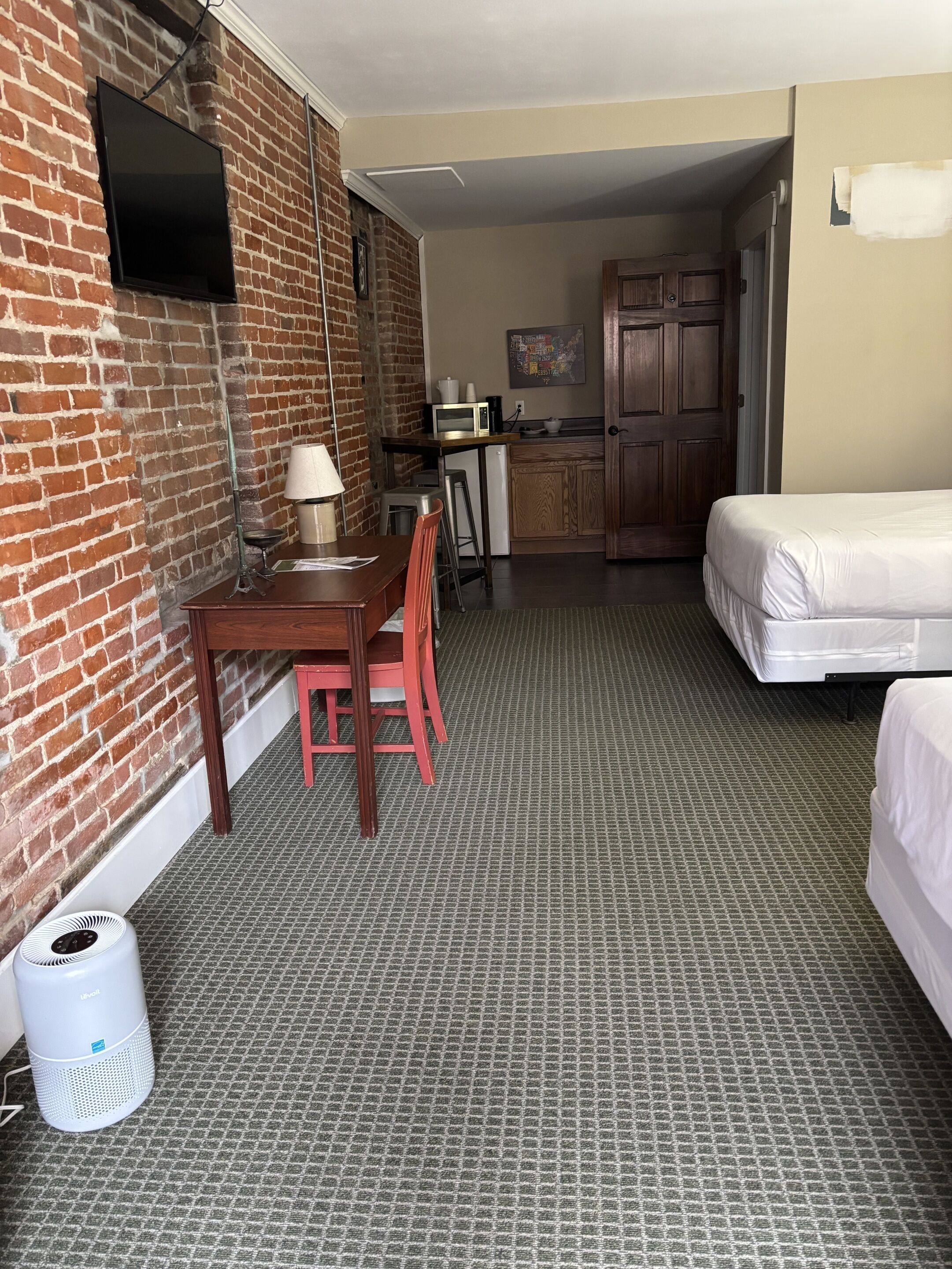 Habitación de hotel con pared de ladrillo, televisor de pantalla plana, mesa de comedor con sillas, kitchenette y cama, además de un suelo con alfombra estampada.