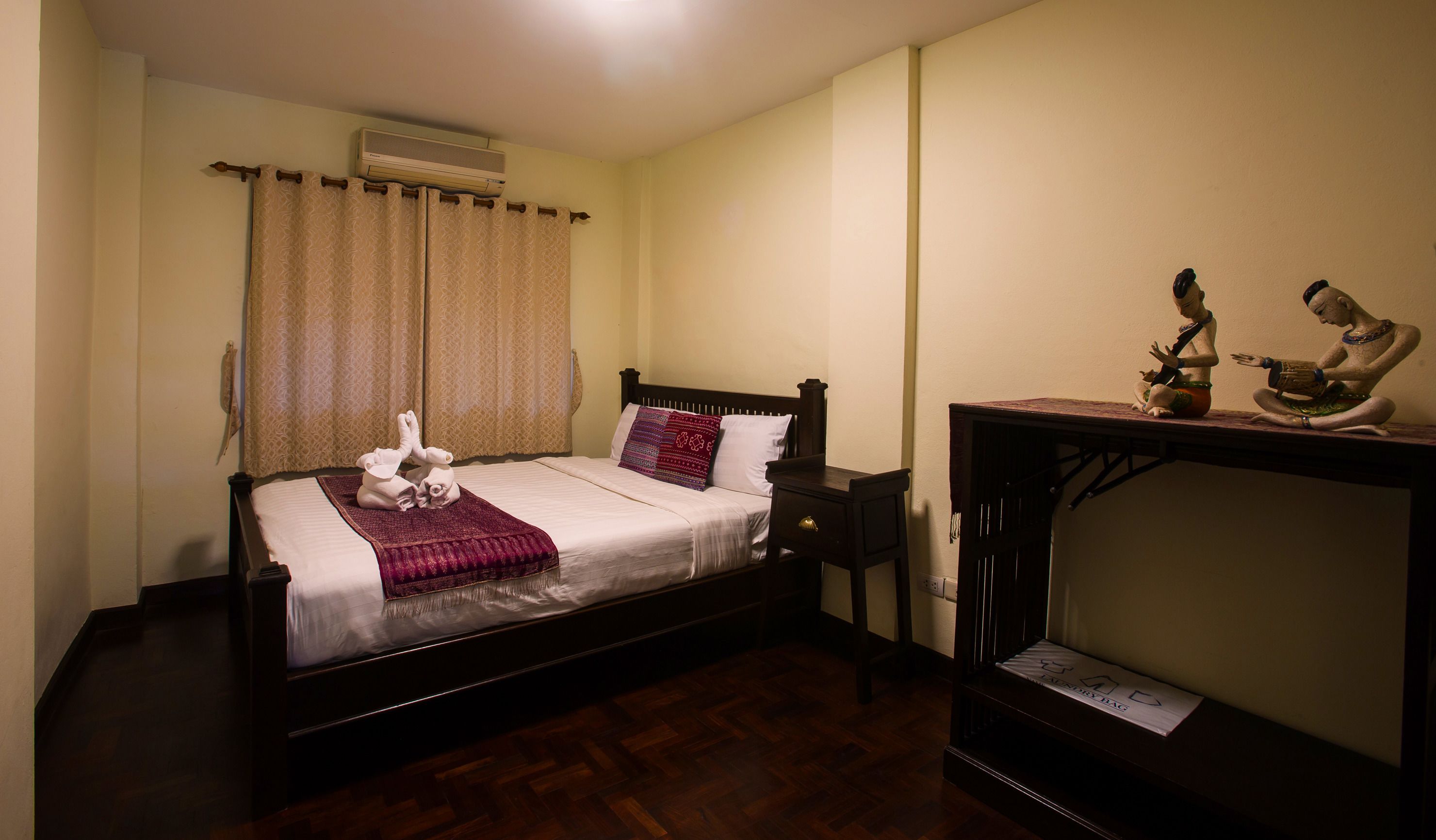 A neatly made bed with a red throw blanket, a wooden nightstand, and a small table with two statues.