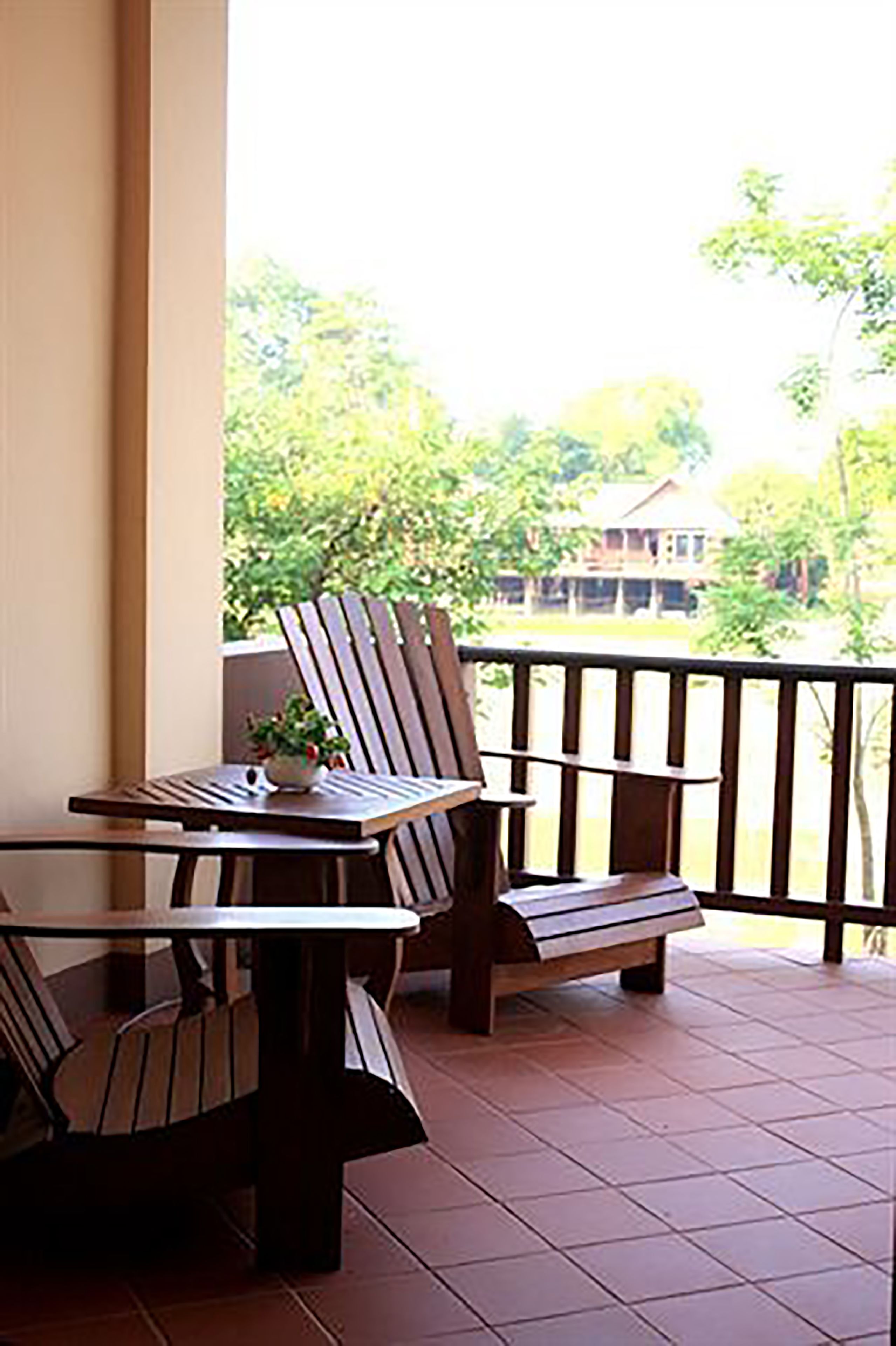 A balcony with a wooden table and chairs, a small plant, and a view of greenery and a building in the background.