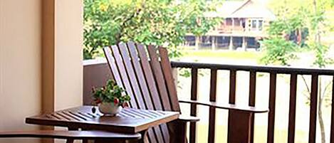 A balcony with a wooden table and chairs, a small plant, and a view of greenery and a building in the background.