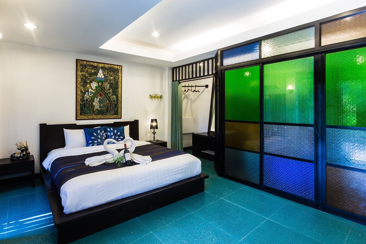 Une chambre d’hôtel moderne avec un grand lit, un élément décoratif mural, une cloison en verre vert et une table de chevet avec une lampe.