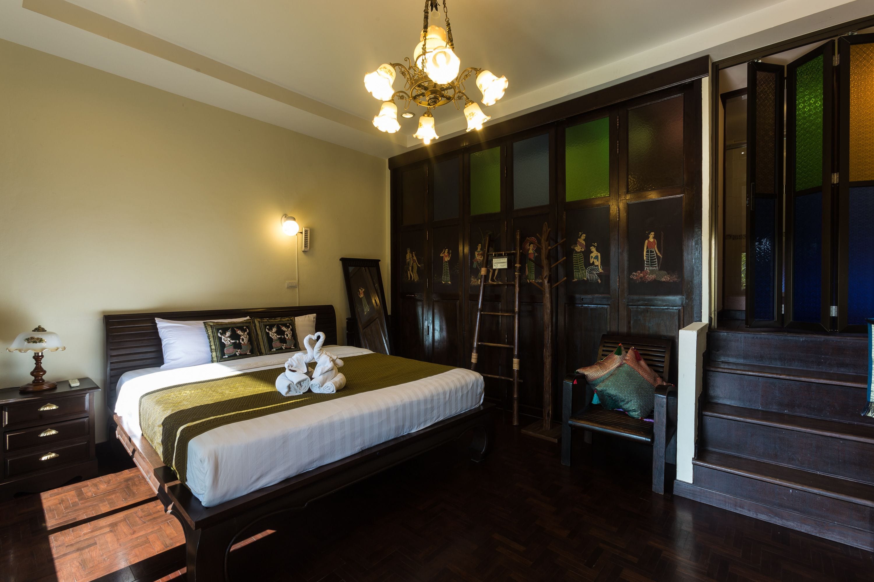 A hotel room with a large bed, a chandelier, a wooden wardrobe, and a bench.