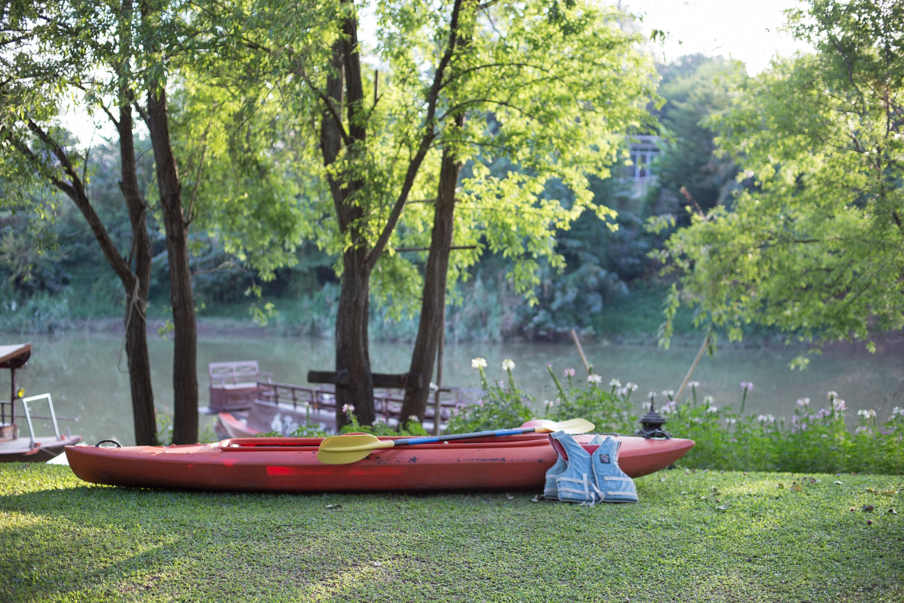 A red canoe with a paddle and a life jacket on a grassy area near a lake.