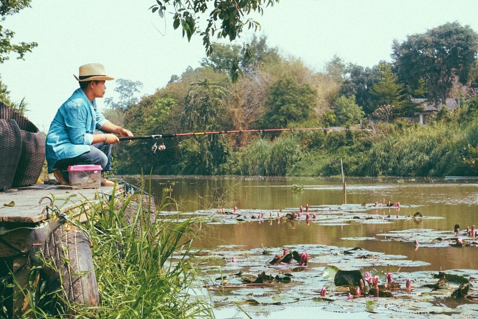 Une personne est en train de pêcher au bord d’une rivière paisible, avec des nénuphars et des arbres en arrière-plan.