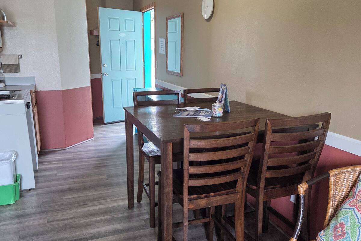 A dining area with a wooden table and chairs, a wicker bench, a kitchen area with a stove, and a door leading to another room.