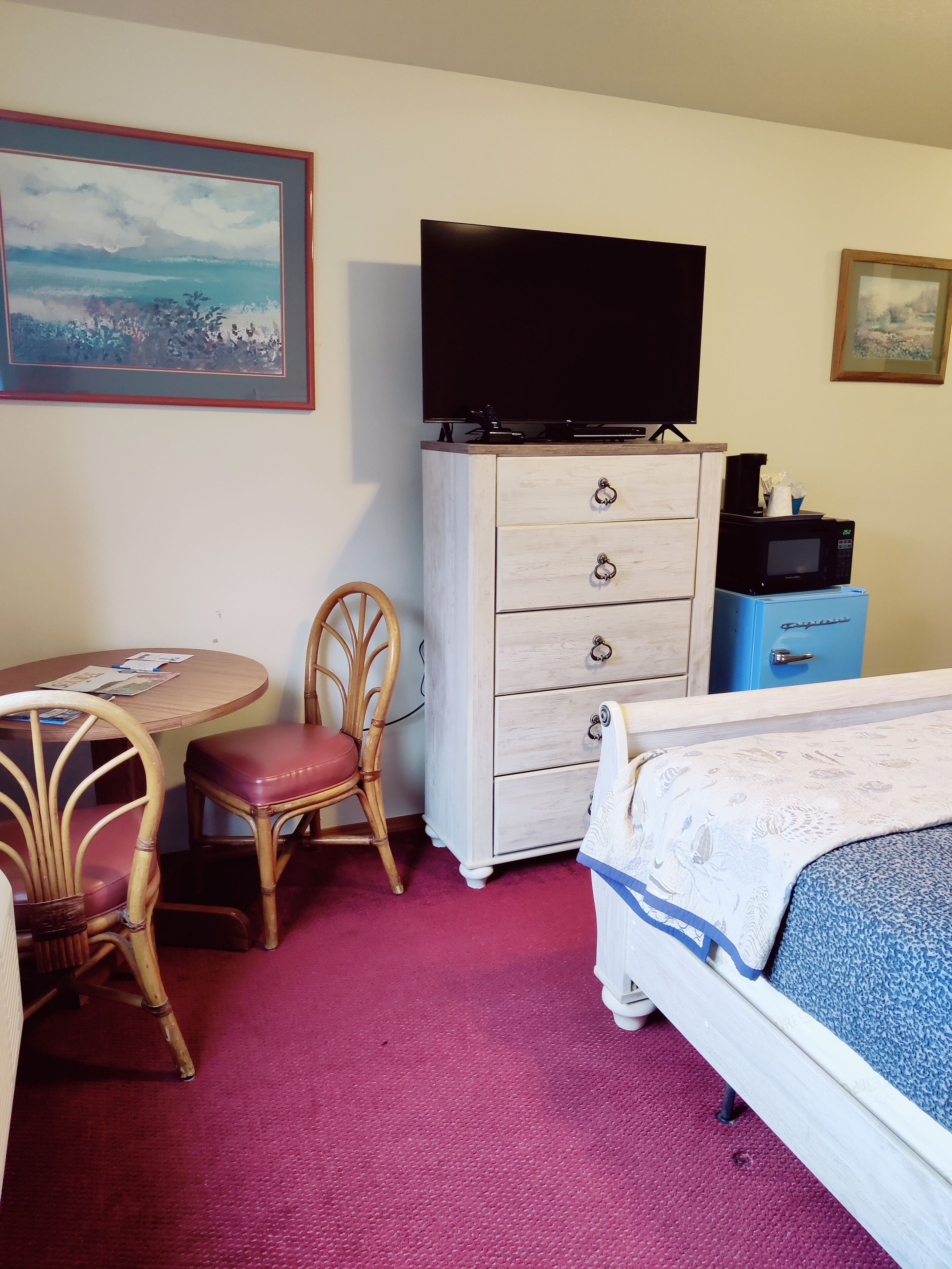 A hotel room with a bed, a desk with a chair, a dresser with a television, and a painting on the wall.