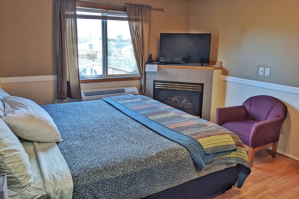 A hotel room with a bed, a chair, a television, a fireplace, and a window with curtains.