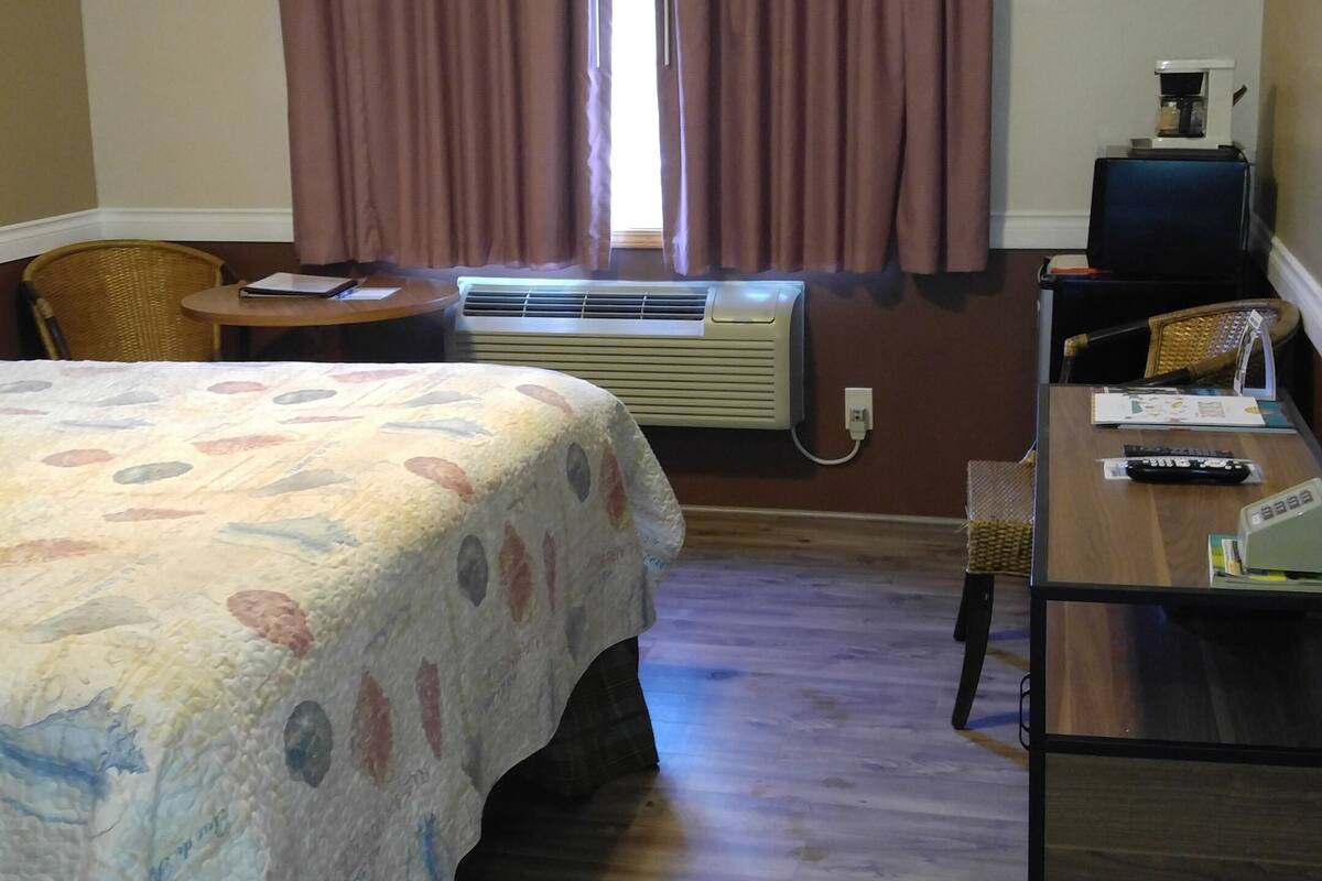 A hotel room with a bed, a desk, a chair, a window with curtains, an air conditioning unit, and a ceiling light.